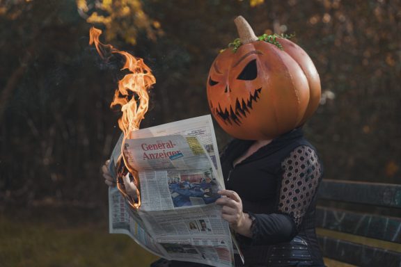 Eine Person mit einem Kürbiskopf liest eine Zeitung, während Flammen aufsteigen.