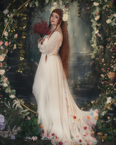 Frau in eleganter, blütenverzierter Robe mit Blumenstrauß vor einer blühenden Kulisse.