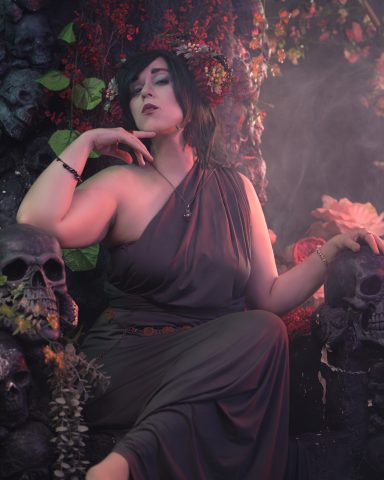 Frau in antikem Kleid mit dunklen Haaren, umgeben von Blumen und Schädeln.