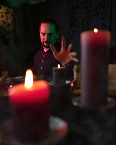 Ein Mann mit erhobener Hand sitzt vor Kerzen und mystischen Gegenständen.