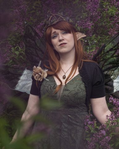 Frau mit roten Haaren und Elfenohren, trägt ein grünes Kleid und schwarze Flügel.