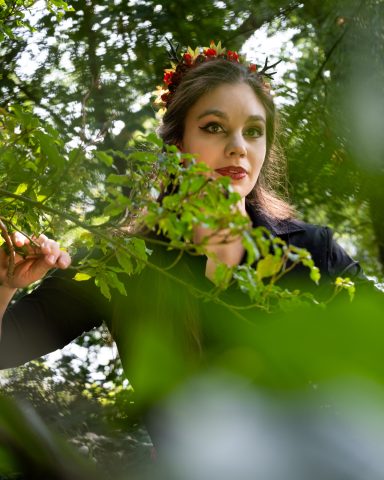 Frau mit Blumenkranz zwischen Blättern im Grünen, schaut aufmerksam.