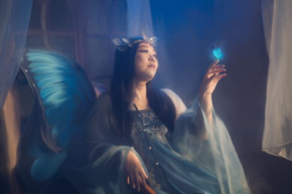 Frau mit Schmetterlingsflügeln hält einen blauen Schmetterling in sanfter Lichtatmosphäre.
