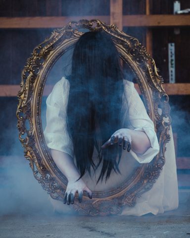 Person mit langen schwarzen Haaren, die aus einem goldenen Spiegel heraus schaut.
