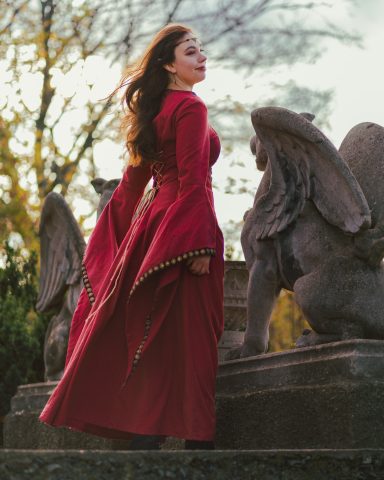 Frau in rotem Kleid steht vor einem monumentalen Löwen, mit wehenden Haaren.