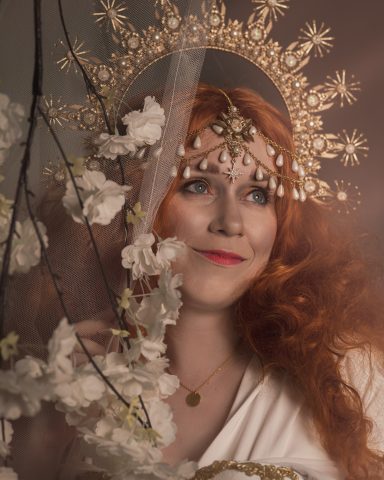 Frau mit rotem Haar, goldenem Kopfputz und floralen Details, lächelnd vor hellem Hintergrund.
