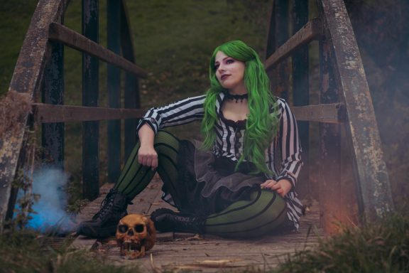 Frau mit grünen Haaren in Streifenoberteil sitzt auf einer Brücke neben einem Totenkopf.